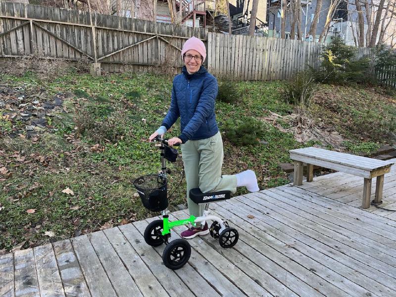 
A white woman wearing a pink toque, blue jacket, and light green pants is posed with her leg on a green and white knee scooter. The leg on the scooter has a cast on it. She is smiling at the camera. Behind her is a yard with green grass but the plants are brown, indicating that it is a cold time of year.
