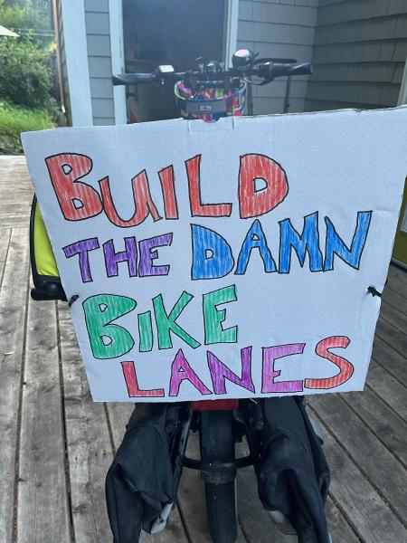 
A cardboard sign attached to the back of a bicycle. It is hand-written in block letters that say "build the damn bike lanes" and coloured with a variety of colours.
