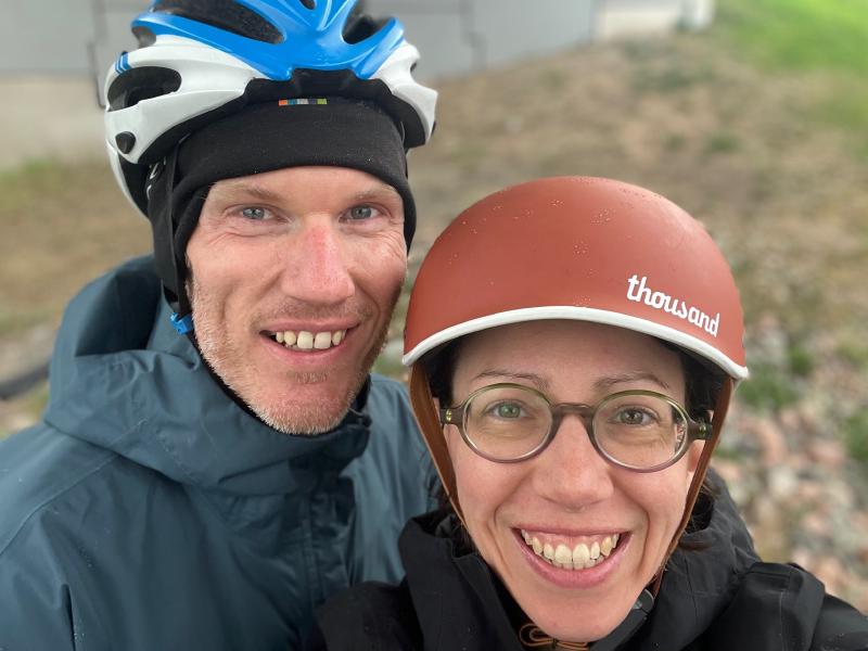 
A selfie of a white woman, wearing glasses and a brown bicycle helmet, along with a white man, also wearing a bicycle helmet. They are smiling at the camera.

