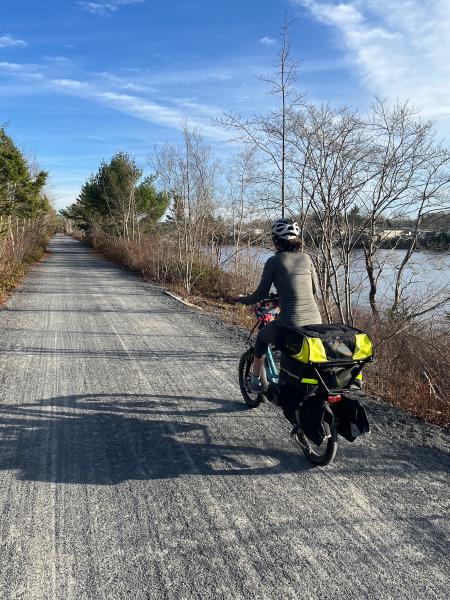 
A woman wearing a gray shirt riding a bicycle away from the camera on a gravel trail. It is a sunny day and her shadow is cast on the trail.
