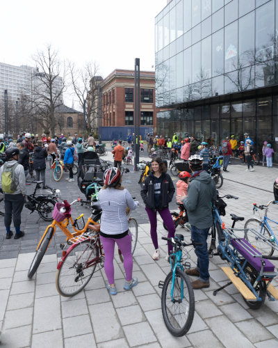 A group of people, including many children, are gathered in a pedestrian plaza. They are outside a large glass building, with brick buildings in the background. Many bicycles, of various shapes and sizes, are also parked in the plaza, among all of the people.