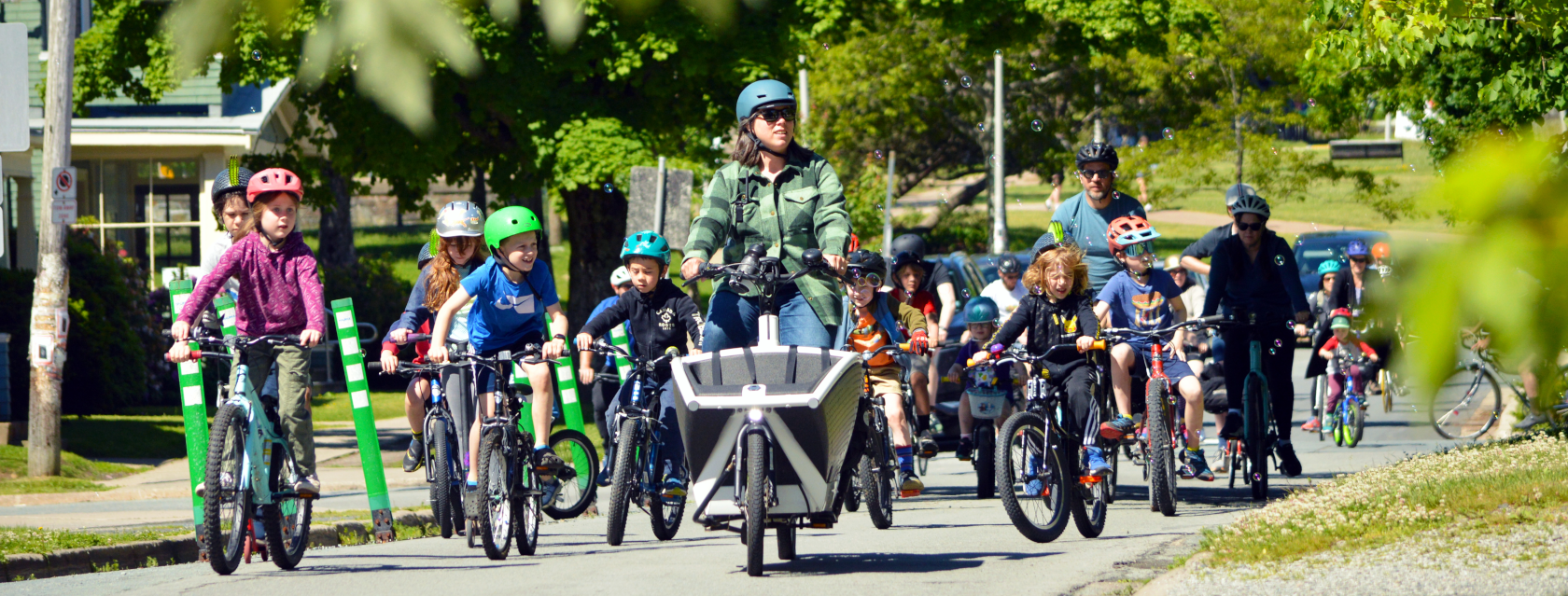 A photo from a Kidical Mass Halifax ride. A group of people are riding bicycles on a tree-lined street. Most of the group in the photo are children on their own bicycles, with a few adults among the group. A woman riding a front-loading cargo bicycle is at the front of the group. The group is so large that it takes up the entire road.