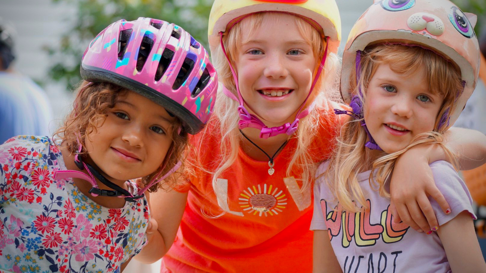 A closeup of three young girls who are leaning in close together and have their arms over each others’ shoulders. The girl on the left has light brown skin, brown curly hair, and is wearing a pink bike helmet and a tshirt with flowers on it. The girl in the middle has white skin, blond hair, and is wearing a yellow bike helmet with a unicorn face and horn on it. Her smile shows that she is missing a few of her front teeth. She is wearing a bright orange tshirt. The girl on the right has white skin, light brown hair and is wearing a light pink bike helmet that has an animal face and cat ears on it. She is wearing a light purple tshirt with some text on it.